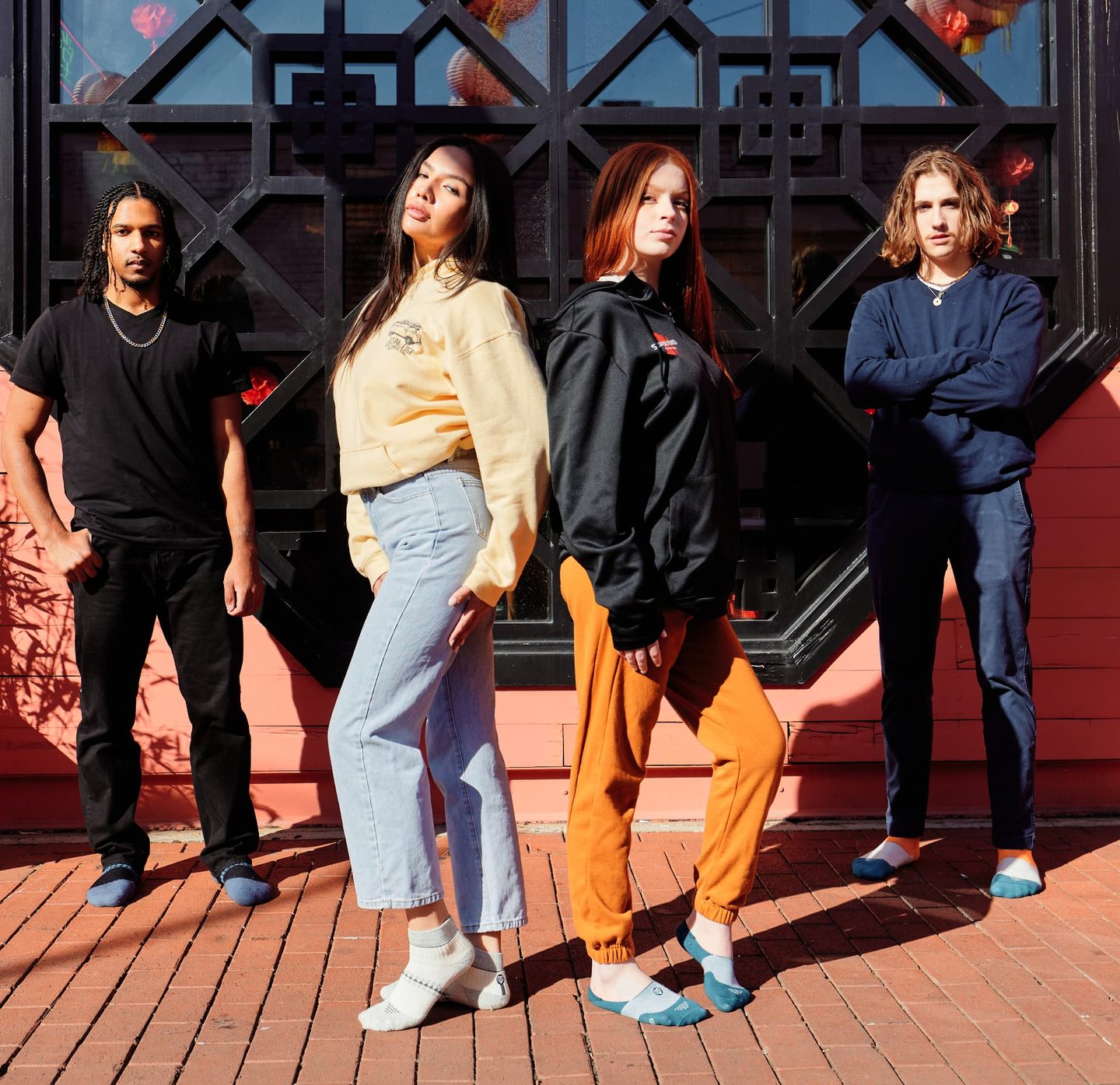 Four models standing in front of a black lattice screen and coral wall in daylight, one wearing a black Super Kind Company hoodie.